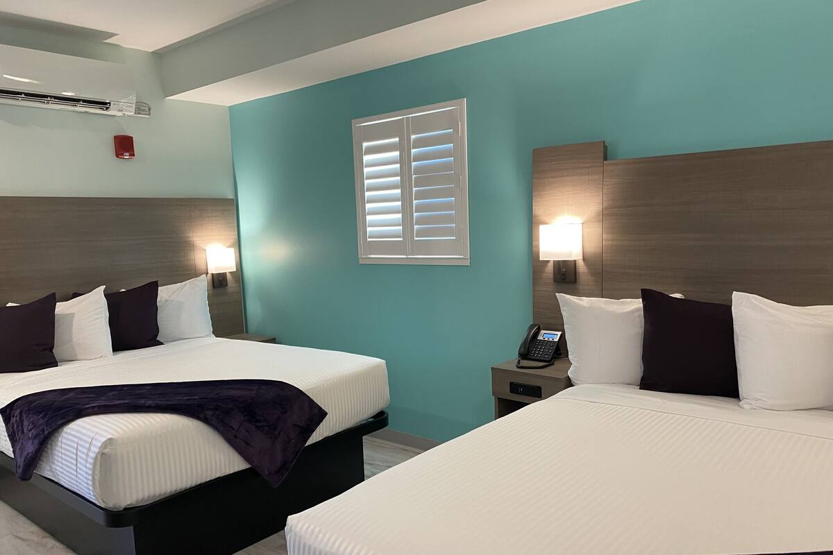 A hotel room with two beds, each with white linens and dark throw blankets, a nightstand with a phone, and a window with blinds.