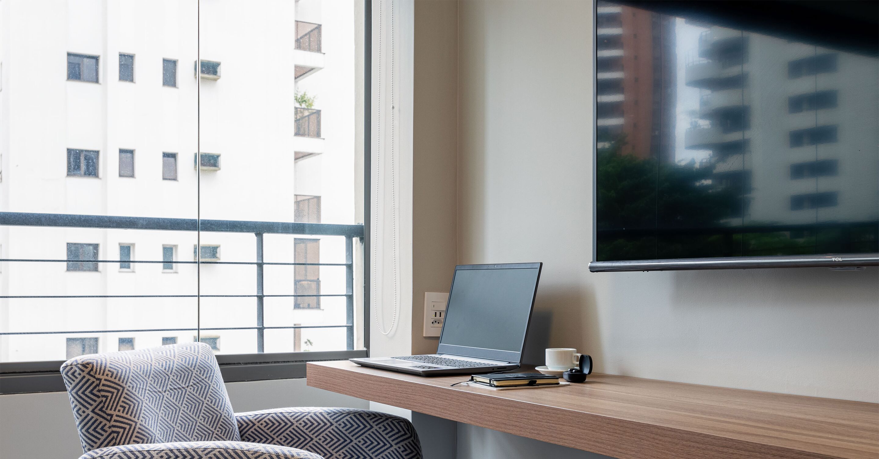 Un espace de travail moderne avec un ordinateur portable posé sur un bureau en bois, une chaise à motifs et une grande fenêtre donnant sur un immeuble.