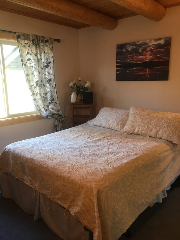 A bedroom with a large bed, a nightstand with a vase of flowers, a painting on the wall, and a window with floral curtains.