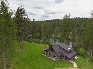 A two-story house with a large deck, surrounded by a forest and a lake.