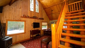 A log cabin interior with a fireplace, wooden walls, a dining area, and a staircase.
