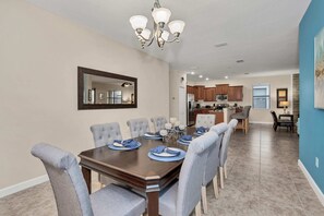 A dining room with a large table set for six, surrounded by grey upholstered chairs. A chandelier hangs from the ceiling, and a mirror is mounted on the wall above the table. - Modern Home, Big Pool Area Near Disney, CDC Cleaning 4cg456 (Davenport)