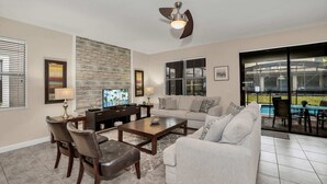 A living room with a television, sofa, coffee table, and chairs. There is a ceiling fan, a fireplace, and a large window with blinds. - Modern Home, Big Pool Area Near Disney, CDC Cleaning 4cg456 (Davenport)