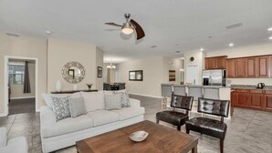 A spacious living room with a sofa, coffee table, and chairs. A kitchen with wooden cabinets and a refrigerator is visible in the background. - Modern Home, Big Pool Area Near Disney, CDC Cleaning 4cg456 (Davenport)