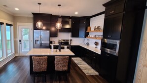 A modern kitchen with dark cabinetry, a central island with bar stools, stainless steel appliances, and pendant lighting. - Emma's View On Lay Lake<br>SUMMERTIME ON LAY LAKE IS THE BEST!!☀️ (Shelby)