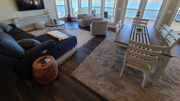 A living room with a dark wood floor, a black leather sofa, a round wicker ottoman, a coffee table, and a dining table with chairs. There are large windows with a view of the water. - Emma's View On Lay Lake<br>SUMMERTIME ON LAY LAKE IS THE BEST!!☀️ (Shelby)