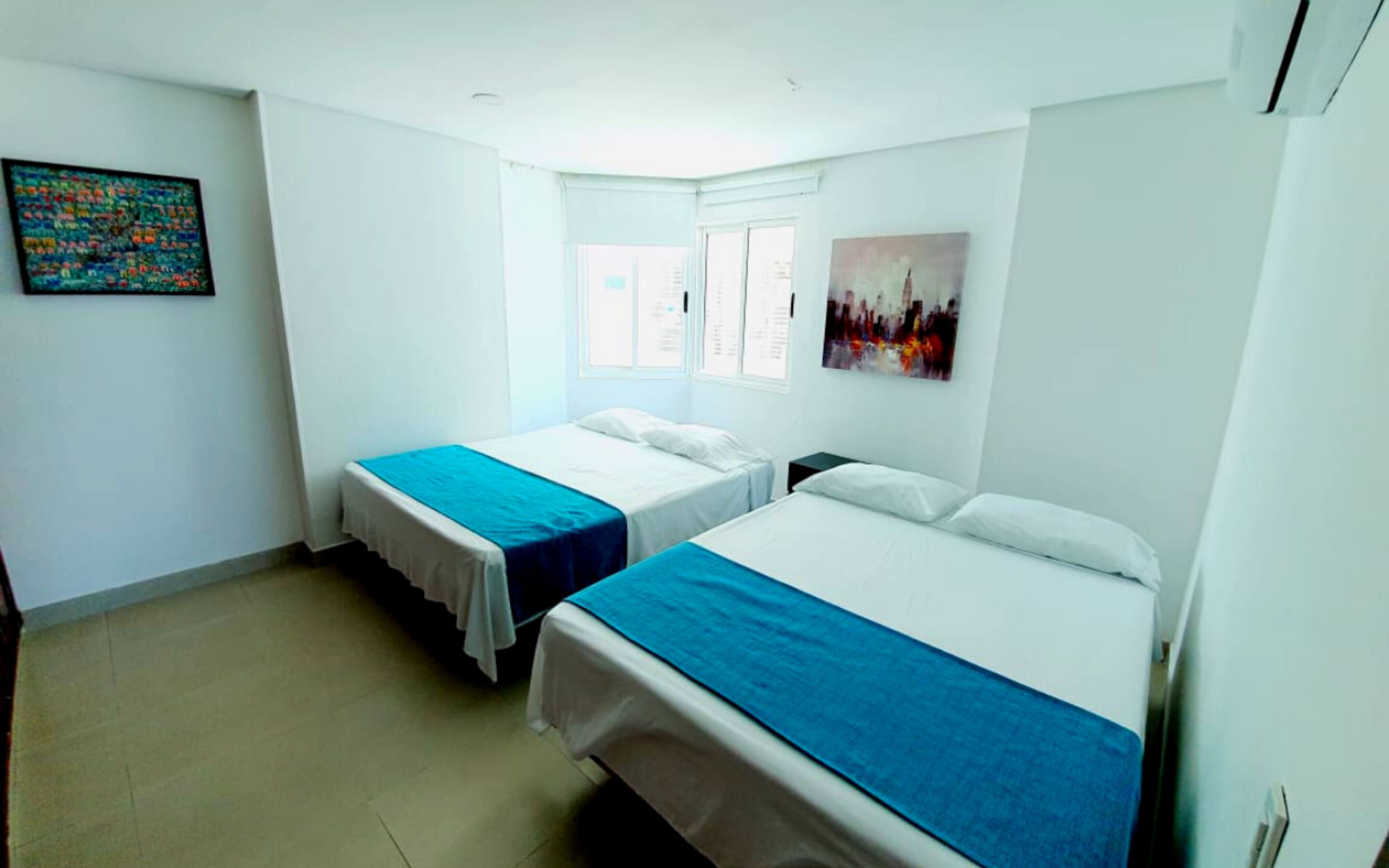 Una camera d'albergo con due letti singoli, una finestra, un quadro appeso al muro e un piccolo tavolino.