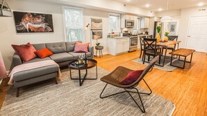 A modern living room with a grey sofa, a coffee table, and a wicker chair. There is a kitchen area with white cabinets and a dining table with chairs. - The Inn at Newton Highlands, boutique apartment-hotel (Newton)