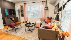 A modern living room with a sofa, armchair, and coffee table, a flat-screen TV, and a large window with blinds. - The Inn at Newton Highlands, boutique apartment-hotel (Newton)
