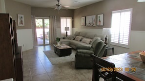A spacious living room with a sectional sofa, a coffee table, and a dining table. There are large windows with blinds, a ceiling fan, and a door leading outside. - We invite you to our Casa in the Sonoran Desert, nestled in San Tan Valley. (San Tan Valley)