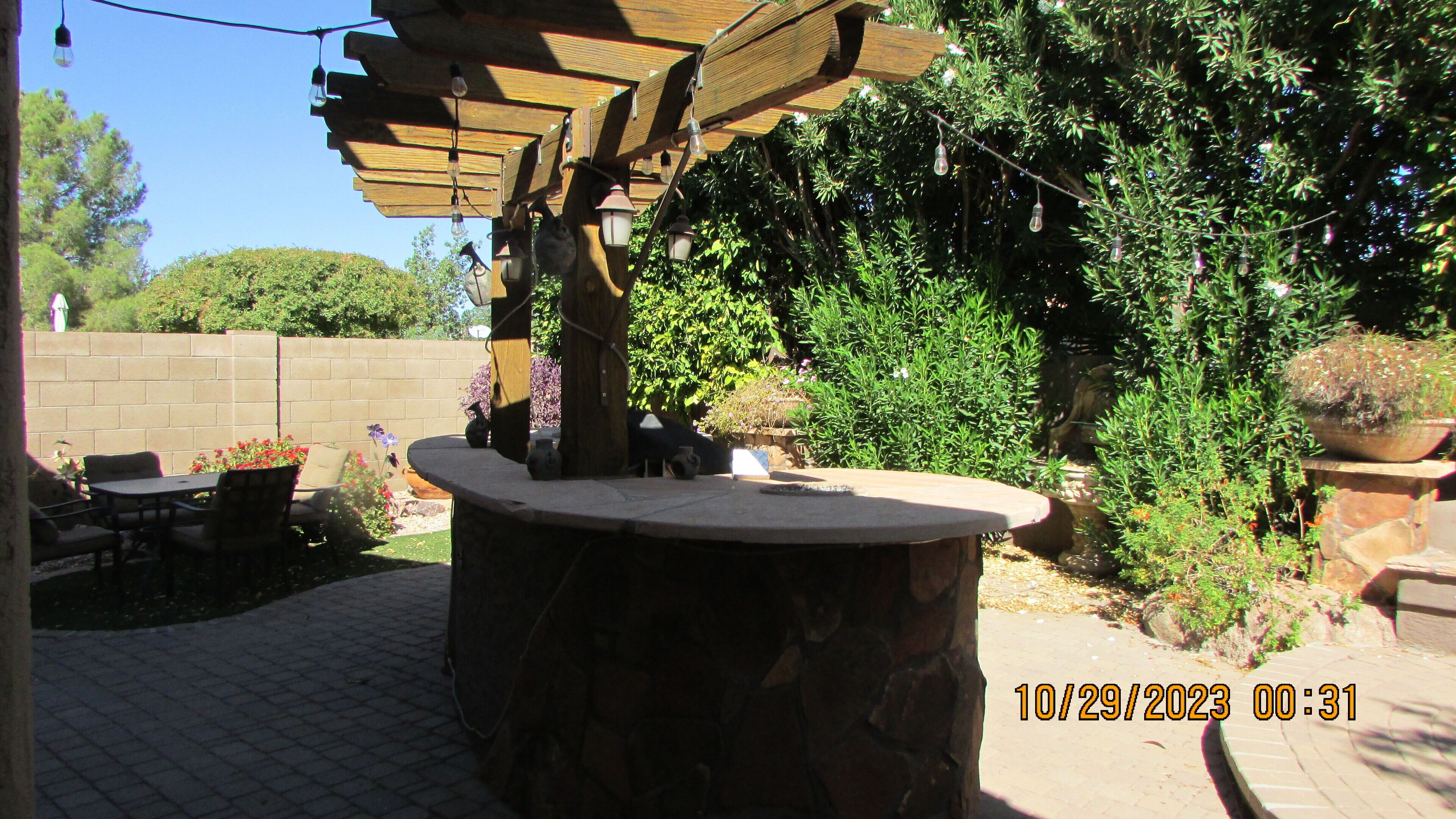 Una mesa de piedra con una pérgola de madera encima, rodeada de plantas en macetas y un muro de ladrillo.