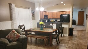 Un comedor con una mesa de madera, sillas y un banco. Se observa una cocina al fondo con armarios de madera y un refrigerador.