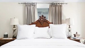 A neatly made bed with white linens and a wooden headboard, flanked by two bedside tables with lamps and a small decorative item. - The Belle at Avery Lafayette Suite (Morganton)