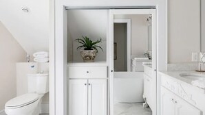 A modern bathroom with a toilet, a cabinet with a plant, a mirror, and a sink. - The Belle at Avery Lafayette Suite (Morganton)