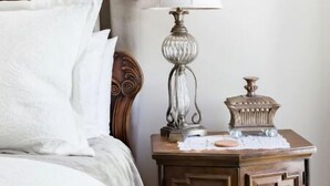 A neatly made bed with white linens, a wooden headboard, a bedside table with a lamp and decorative items, and a window with curtains. - The Belle at Avery Lafayette Suite (Morganton)