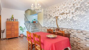 Une salle à manger avec une nappe rouge, des chaises en bois et un lustre. Un escalier orné d’une rampe décorative mène à un niveau supérieur.
