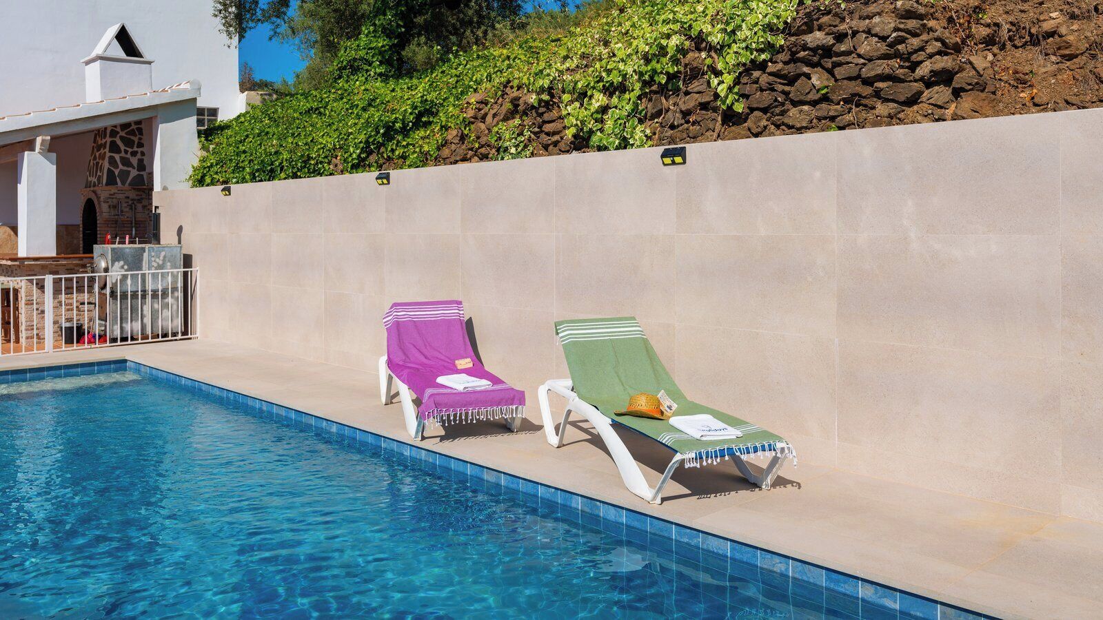 Une piscine avec deux chaises longues, l’une recouverte d’un tissu violet et l’autre d’un tissu vert, et un mur de pierre avec de la verdure derrière.