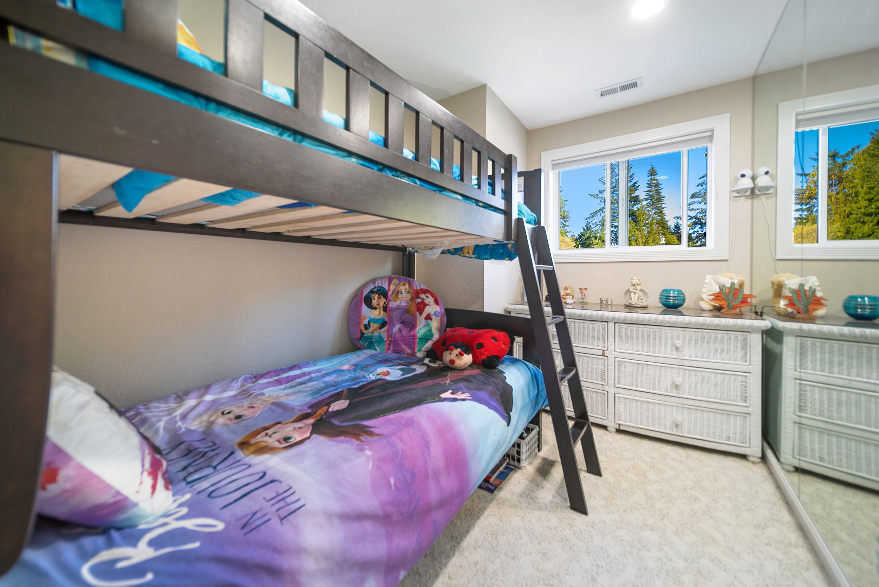 A bunk bed with a Disney-themed bedspread, a ladder, a dresser, and a window with a view of trees.