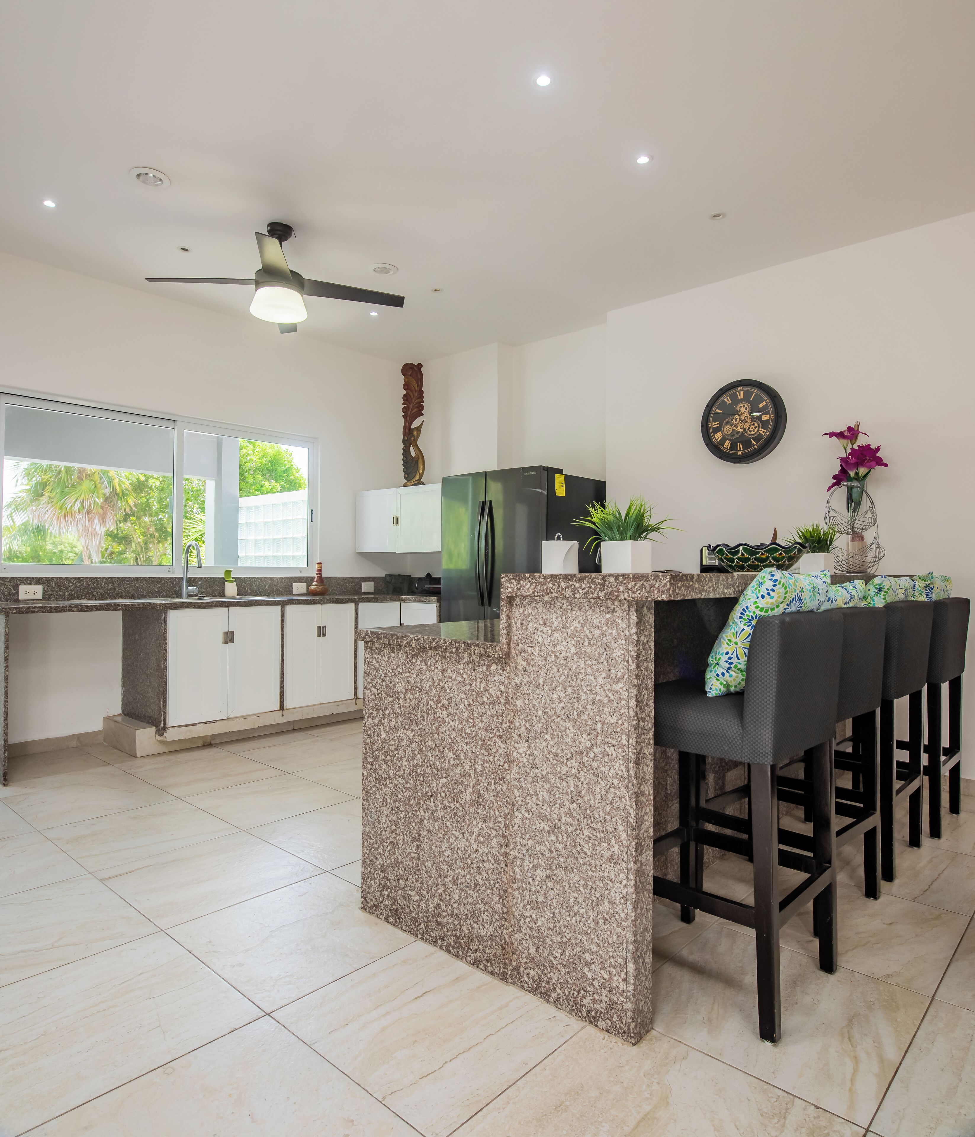 A modern kitchen with granite countertops, a built-in refrigerator, white cabinets, a dining area with black bar stools, and a ceiling fan.