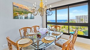 A dining area with a glass table, wooden chairs, a pineapple-themed chandelier, and a large window with a view of the ocean and buildings.