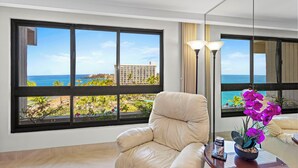 A room with a large window offering a view of the ocean and a building. There is a beige armchair and a small table with a plant and a can of beer.