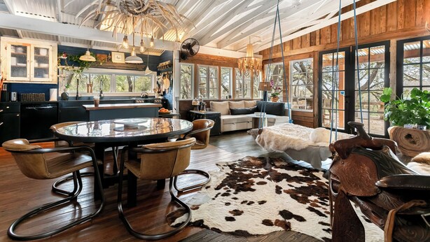 Un vaste espace de vie comprenant une table à manger ronde, des fauteuils en cuir, un tapis en peau de vache et une grande fenêtre donnant sur l’extérieur.