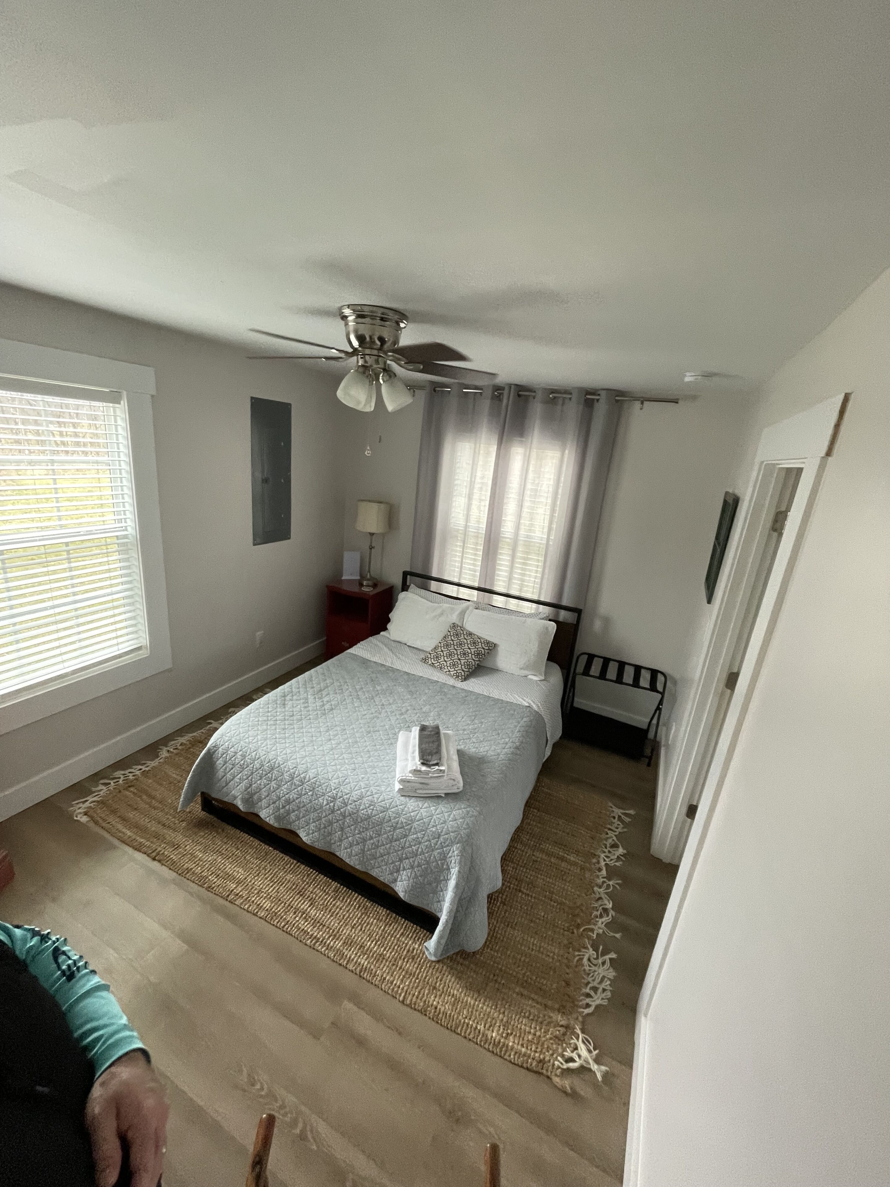 Un dormitorio con cama, ventilador de techo, mesita de noche, lámpara y ventana con persianas.