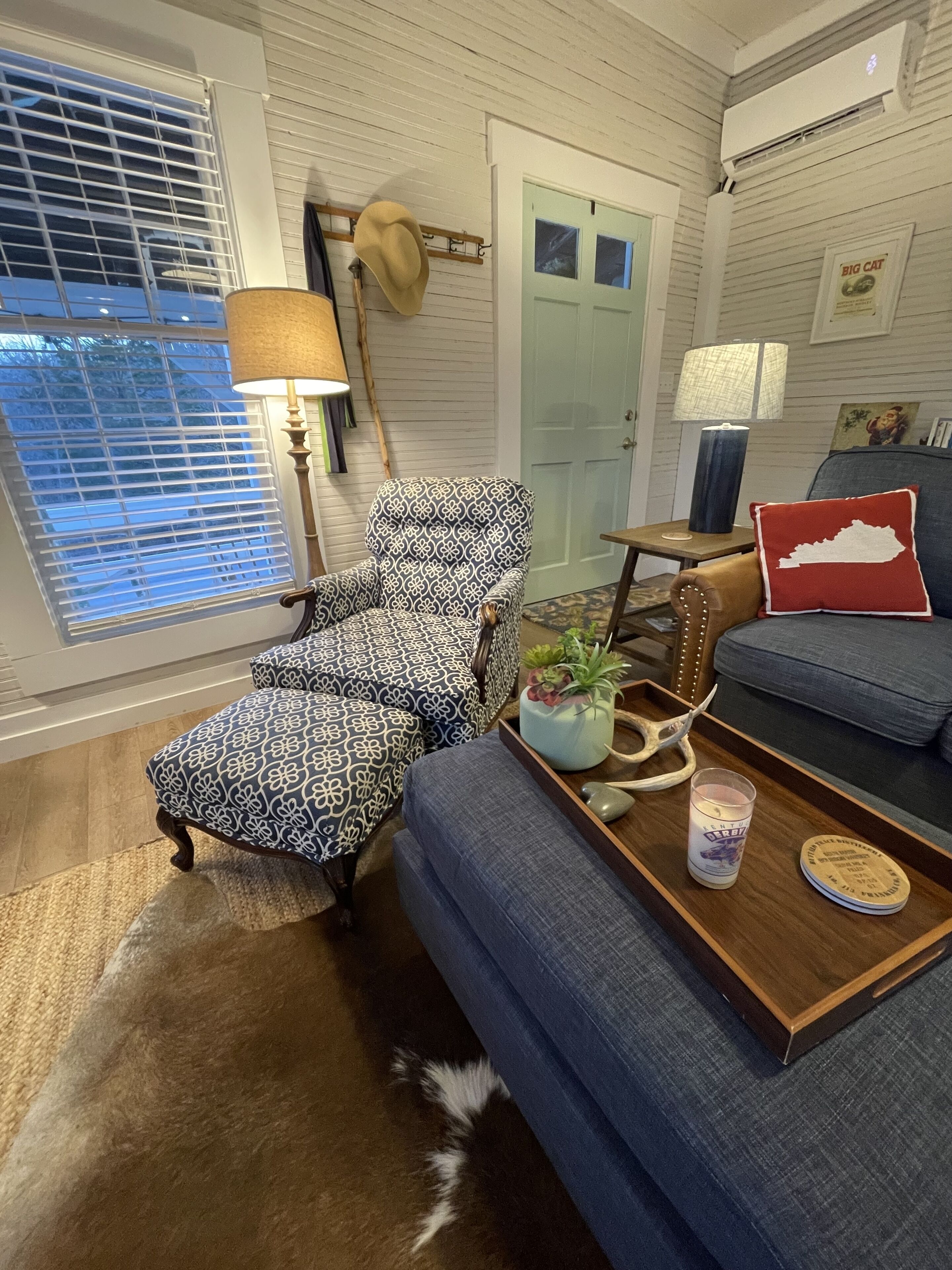 Una sala de estar acogedora con un sillón estampado, un sofá azul, una mesa de centro de madera con una vela y una lámpara.