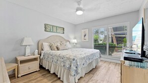 Un dormitorio con cama, mesitas de noche, ventilador de techo, ventana con persianas y vista a zonas verdes.