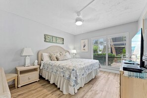 A bedroom with a bed, bedside tables, a ceiling fan, a window with blinds, and a view of greenery outside.