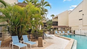 Zona de piscina con sillones reclinables, palmeras y un edificio con balcones.