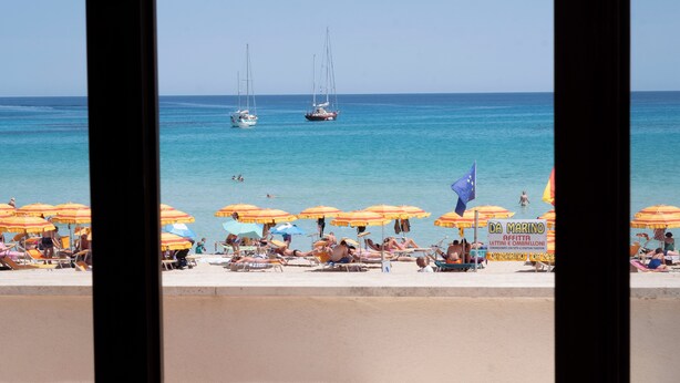 Vista da praia com vários veleiros, guarda-sóis e cadeiras de praia.
