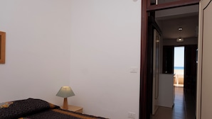 Quarto com cama, mesa de cabeceira com abajur e vista para a praia através de uma porta aberta.