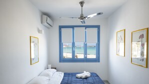 A bedroom with a bed, a ceiling fan, an air conditioning unit, a window with a view of the sea, and framed pictures on the wall. - Casa Blu, a house directly by the sea and in a central location in San Vito Lo Capo. (SAN VITO LO CAPO)