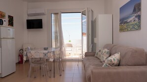 Una sala de estar con un sofá color crema, una mesa de comedor con sillas, un televisor montado en la pared y una puerta que da a un balcón.