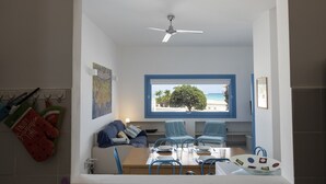Comedor con una mesa preparada para cuatro personas, un ventilador de techo y una ventana con vista a la playa.
