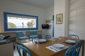 Comedor con mesa de madera dispuesta para cuatro personas, sillas azules y un ventanal con vista al mar.