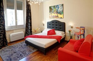 A bedroom with a bed, a red sofa, a small table with a vase, a painting on the wall, and a window with blinds. - HQH Trevi (Rome)