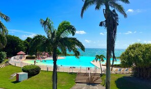 Ein Poolbereich mit klarem, blauem Wasser, umgeben von Palmen und Blick aufs Meer.
