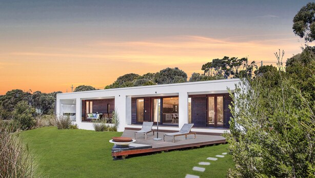 A modern two-story house with a flat roof, large glass windows, and a deck with outdoor furniture. - Secluded Luxury (Greens Beach)