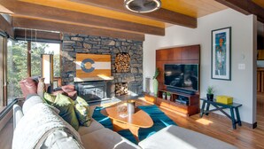 Un salon moderne doté d’une cheminée en pierre, de poutres apparentes en bois, d’un grand téléviseur et d’un coin salon confortable.