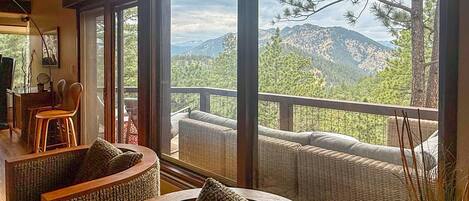 Una habitación con un amplio ventanal que ofrece vistas a un paisaje montañoso, amueblada con sofás de mimbre y una silla de madera.