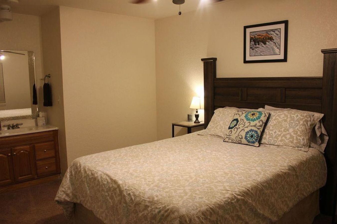 A bedroom with a bed, a nightstand, a lamp, a mirror, and a framed picture on the wall.