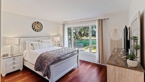 Ein Schlafzimmer mit einem Bett, Nachttischen, einer Uhr, einem Fenster mit Vorhängen und Blick auf eine grüne Landschaft.