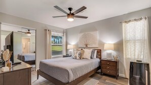 Un dormitorio con cama, mesitas de noche, un espejo, ventilador de techo, ventana con cortinas y vistas al exterior.