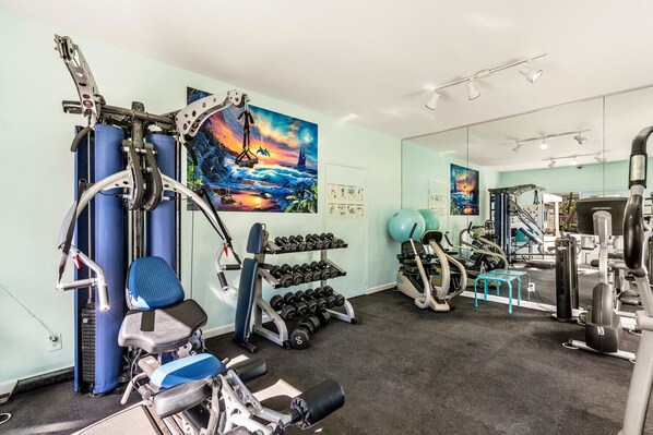 A gym with exercise equipment, a blue chair, and a painting of a sunset. - Fairway Terrace F-107: Waikoloa Condo Golf Course View With Pool (Waikoloa)
