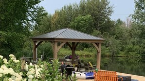 A gazebo with outdoor seating, surrounded by lush greenery and a pond.