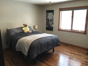 A bedroom with a large bed, a teddy bear, a painting, a window, and a lamp.