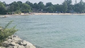 A beach with rocky shoreline, calm water, and a distant shoreline with buildings and trees.