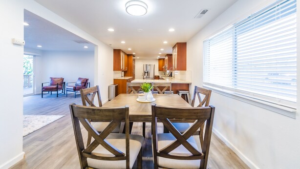 A modern dining area with a wooden table and chairs, a kitchen in the background, and a large window with blinds.